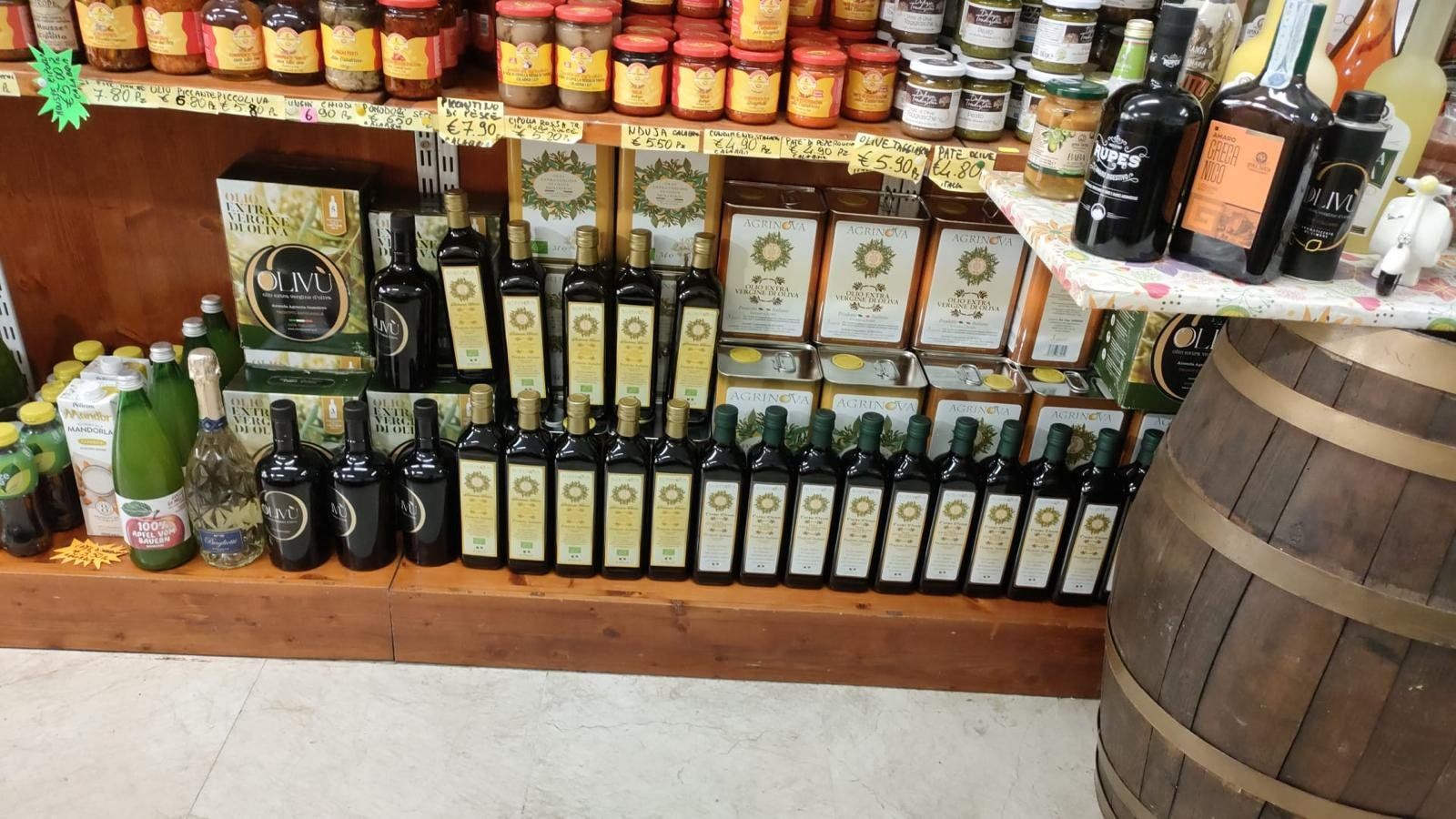 Il banco dell’olio extravergine di Natural Fruit: le file di bottiglie con l’etichetta verde e oro, i cartoni OLIVÙ e le confezioni Agrinova sullo scaffale di legno, con la botte accanto